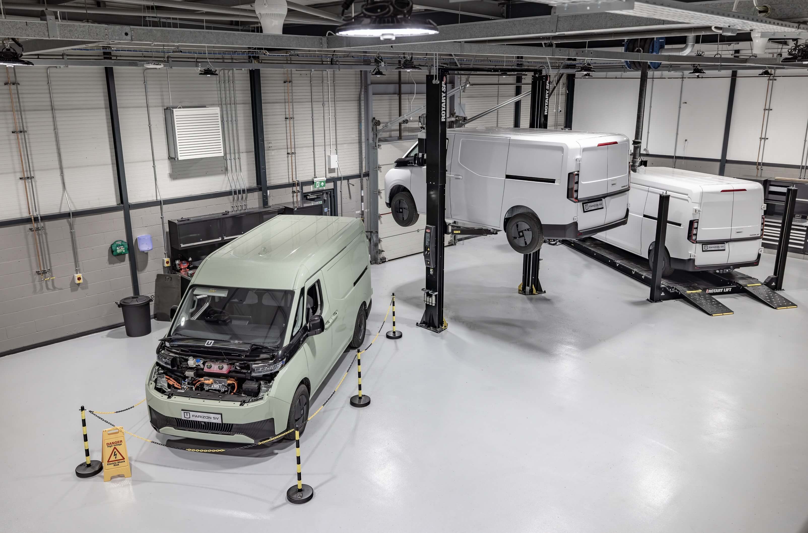 Electric vans in a clean automotive workshop, one with an open hood and two lifted on hydraulic service platforms.
